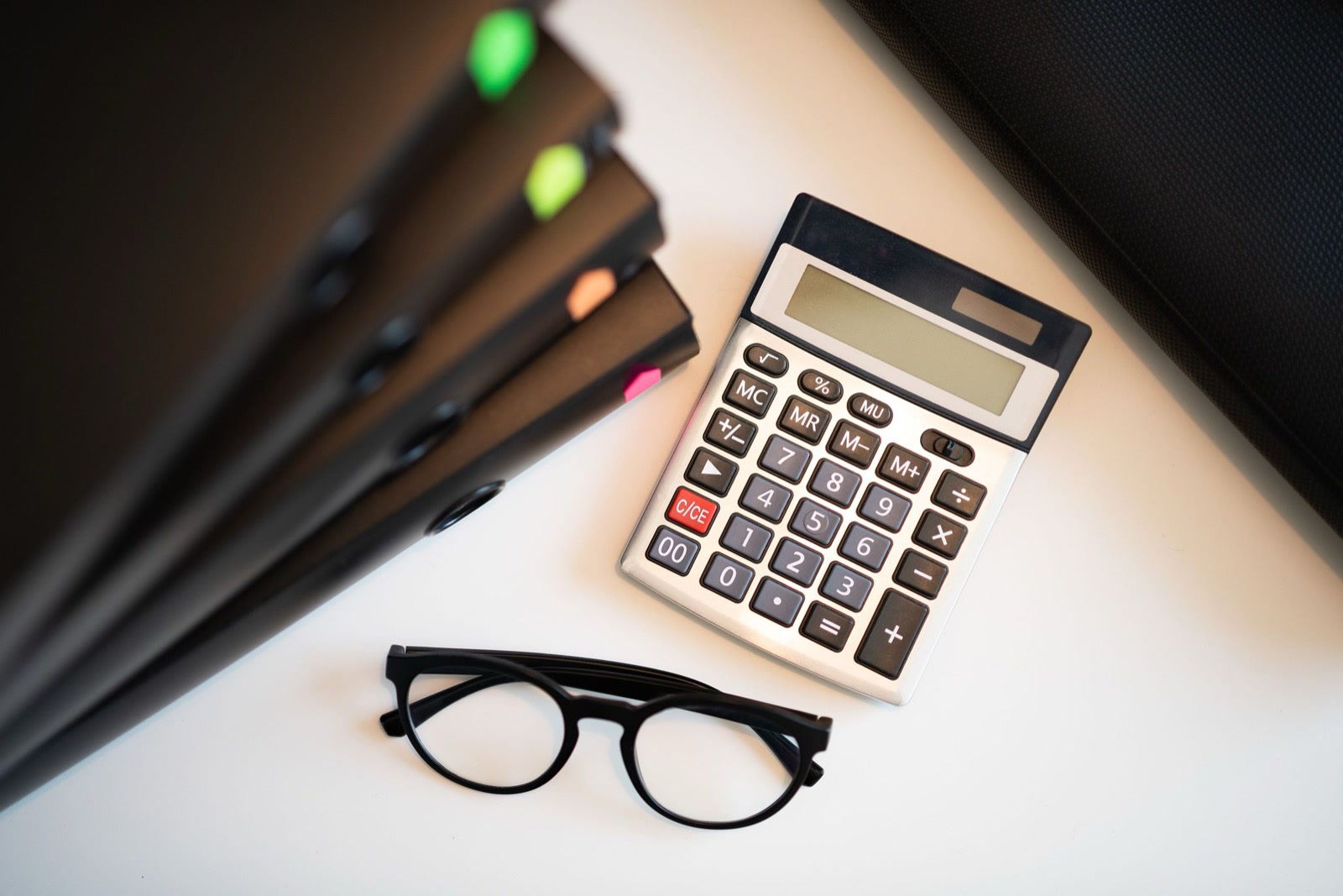 A desk calculator, a pair of black-framed glasses and a row of black ring binders with coloured tabs on a white desk, seen from above