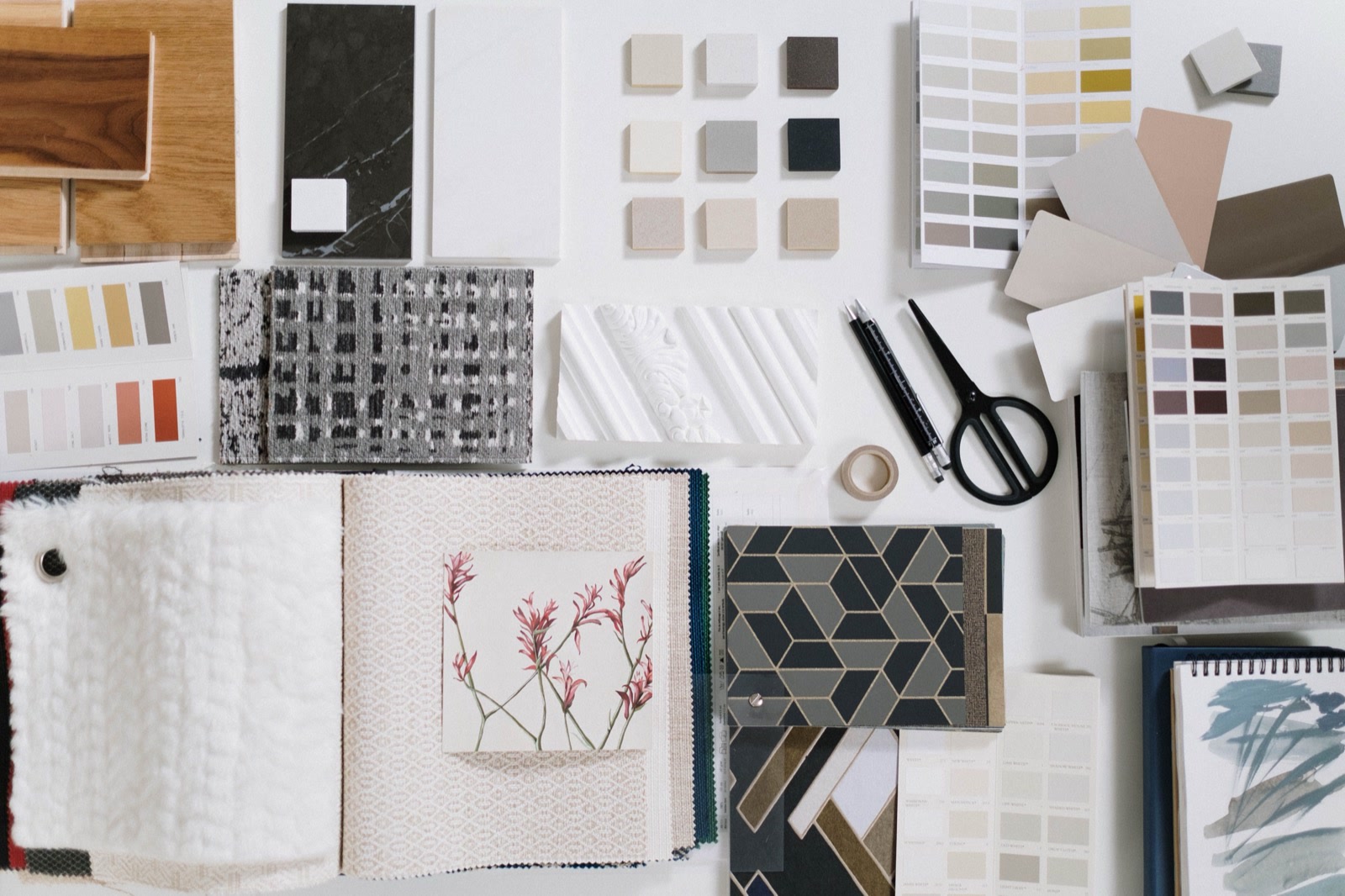 A flat lay of interior design samples on a white table: wood flooring, stone and tile pieces, paint colour charts, fabric books and wallpaper