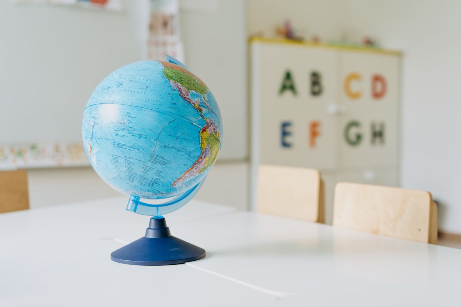 A desk globe on a white table in a classroom, with wooden chairs and a cupboard marked with letters of the alphabet behind it