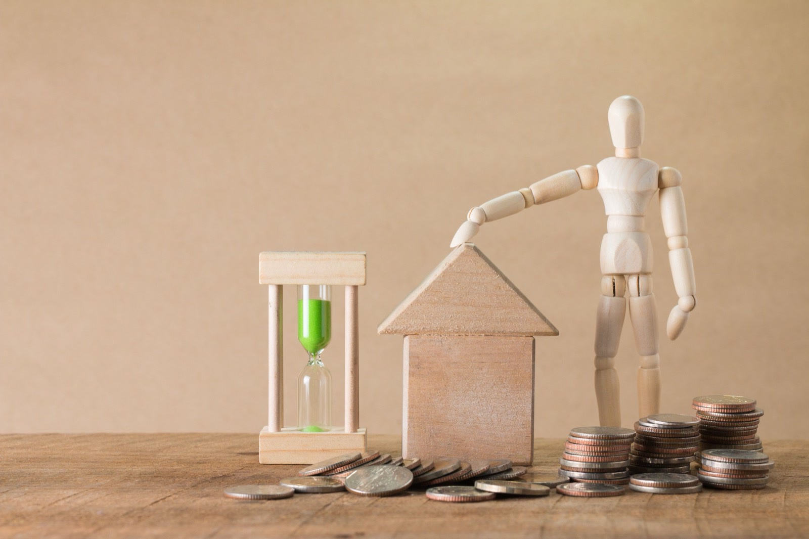 A wooden model house beside stacks of coins and an hourglass, with a wooden artist's figure resting a hand on the roof