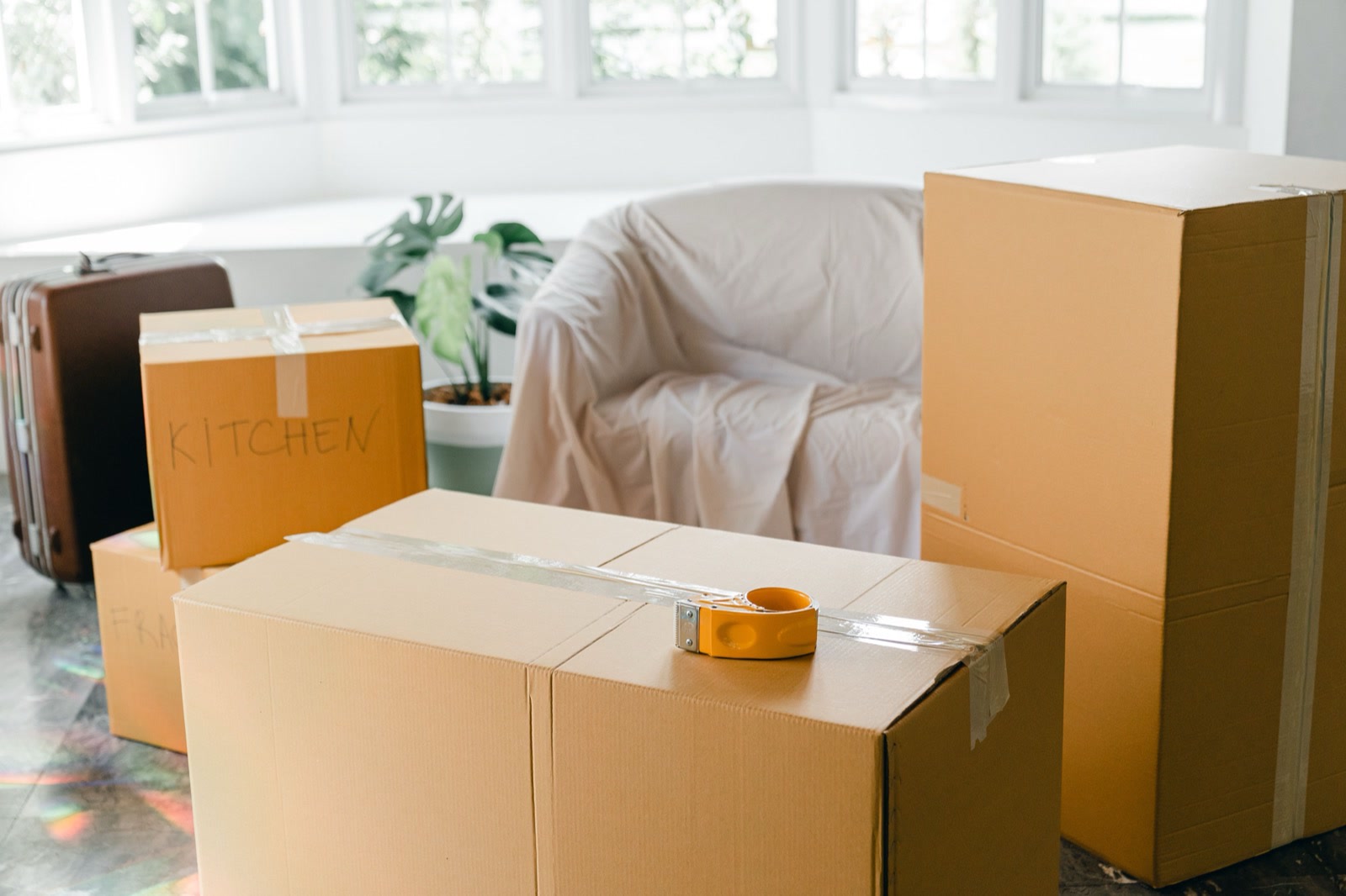 Sealed cardboard moving boxes, one marked for the kitchen, with a tape dispenser, a suitcase and an armchair under a dust sheet in a bright room