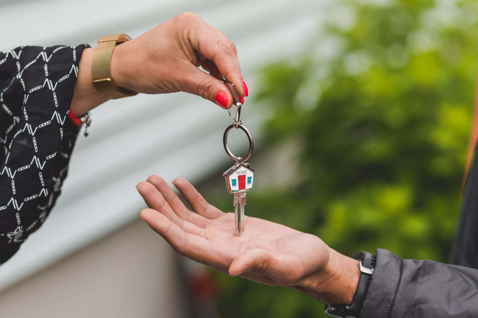 One hand passing a house-shaped key on a key ring into the open palm of another hand, outside a home