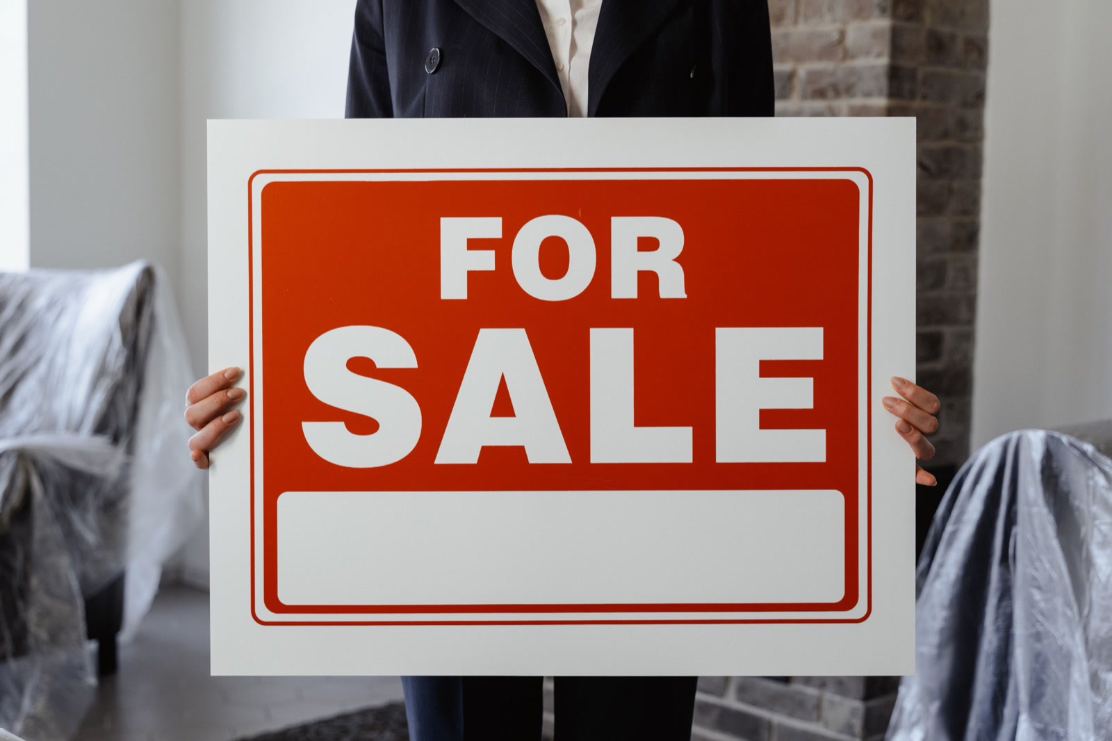 A person in a dark suit, seen from the shoulders down, holding a red and white For Sale sign in a furnished room