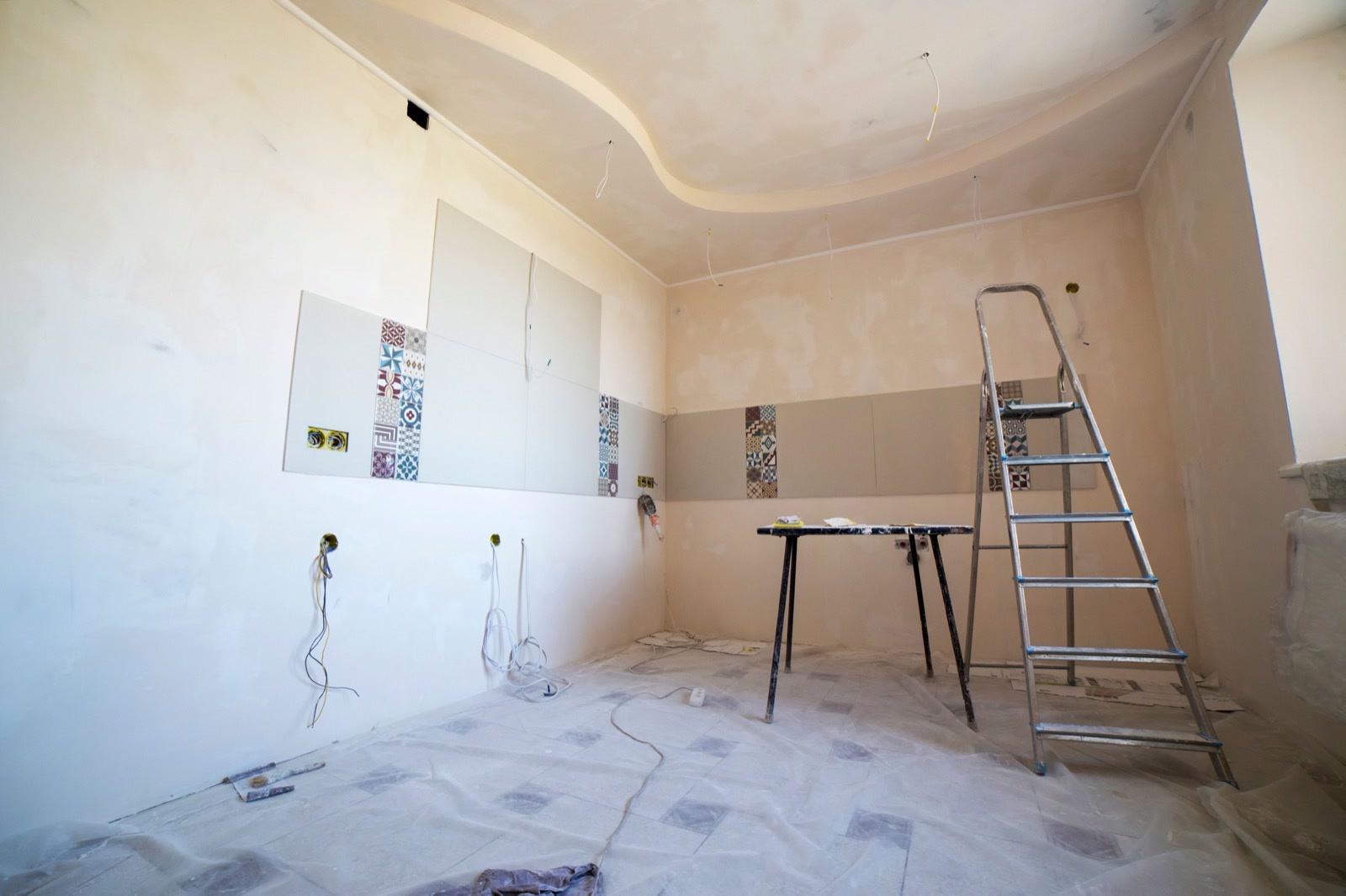 A room in the middle of renovation, with wall tiles partly laid, loose wiring, a stepladder and dust sheets on the floor